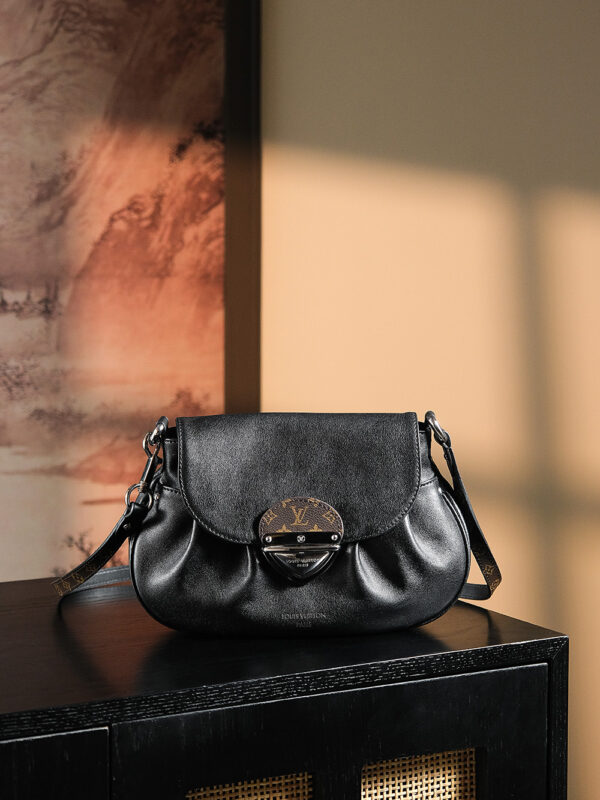 Black leather saddle bag with metal buckle, brand emblem, adjustable strap, placed on black wooden furniture